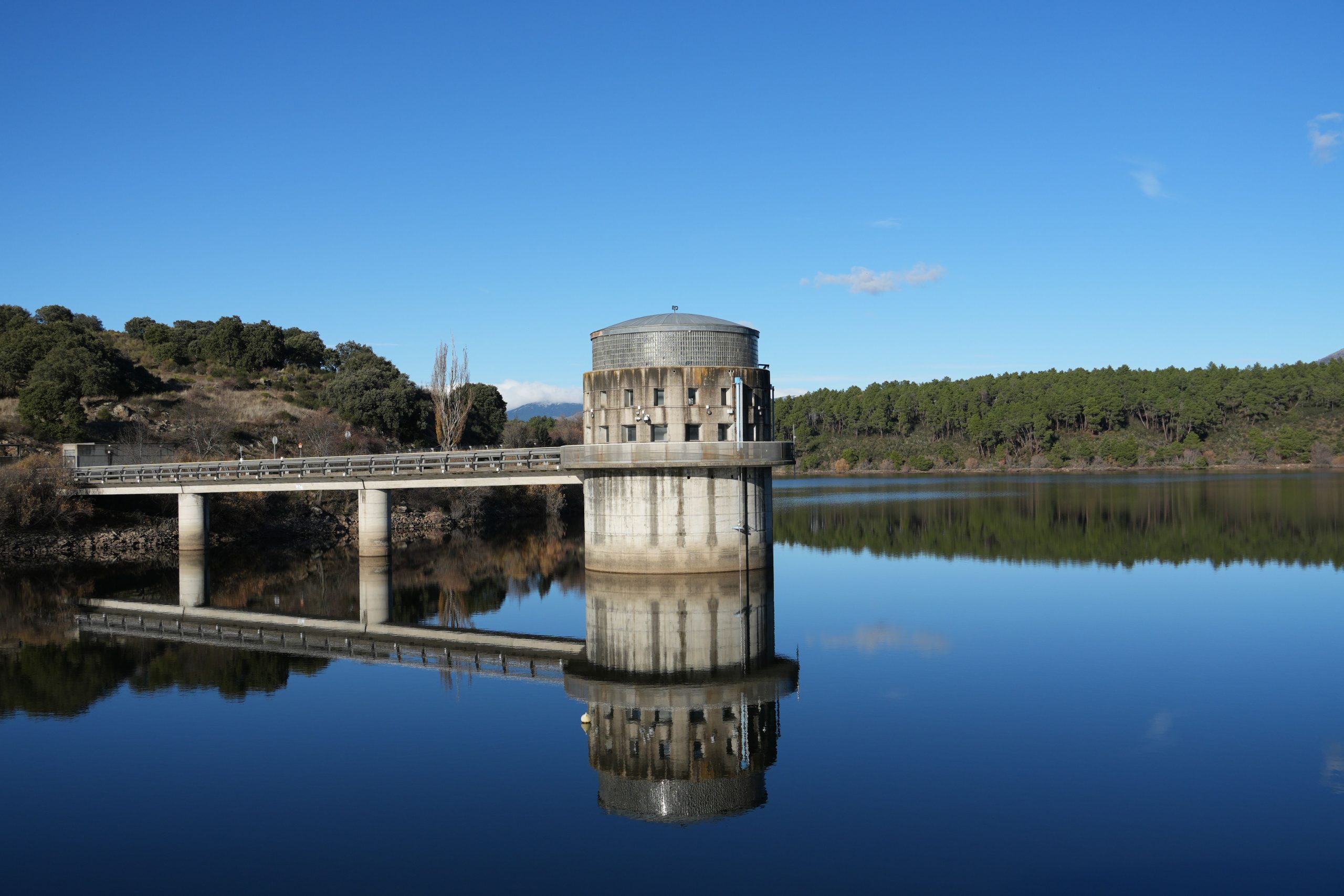Represa o sala de control rodeada de agua calmada y paisaje boscoso bajo cielo azul.